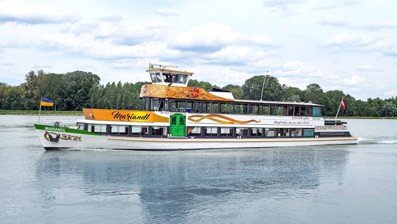 MS Mariandl, © Art & Industrial Design Ein Ausflugsschiff namens MS Mariandl fährt auf einem Fluss, umgeben von Bäumen. Es trägt eine ukrainische und eine österreichische Flagge.