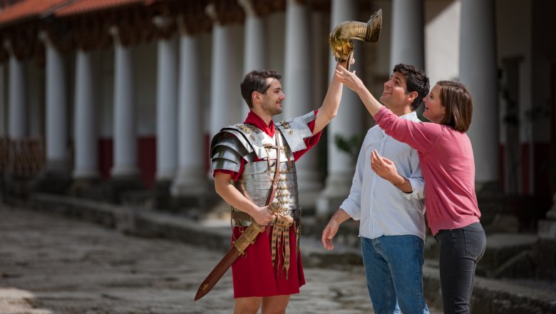 Cäsarentrophy, © Atelier Olschinsky Ein Mann in römischer Rüstung hält einen Helm, während zwei Personen ihn bewundern.