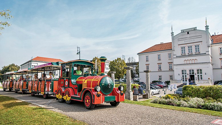 Ein bunter Touristenzug fährt an einem weißen Gebäude mit der Aufschrift 'Rathaus' vorbei.
