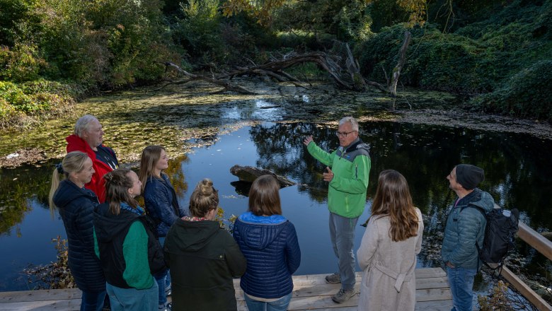 Eine Gruppe von Menschen steht auf einem Holzsteg vor einem Teich in einem bewaldeten Gebiet. Ein Mann in grüner Jacke spricht zu ihnen.