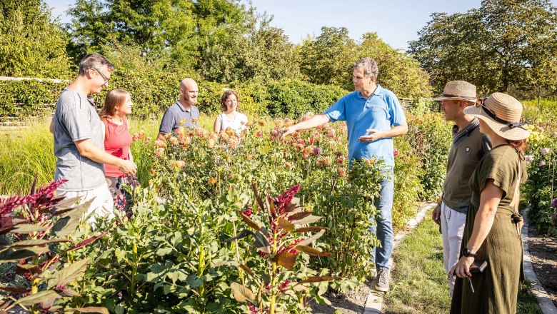 Eine Gruppe von Menschen steht in einem blühenden Garten und hört einem Mann zu, der etwas erklärt.