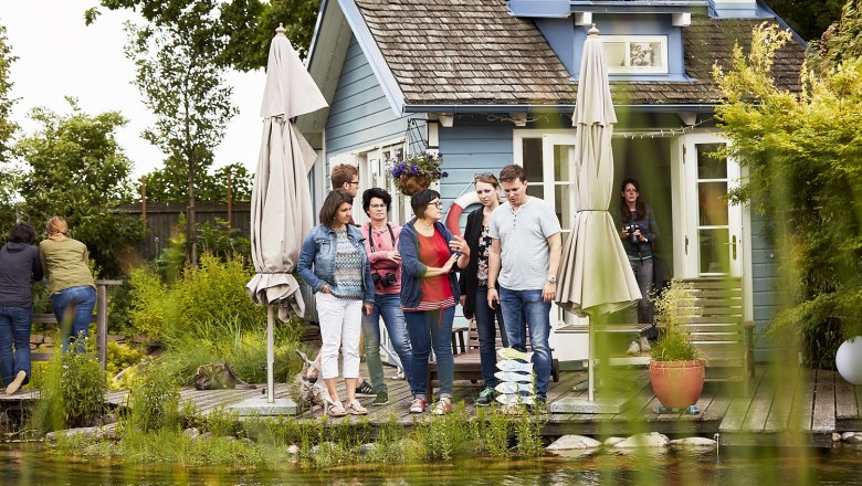 Eine Gruppe von Menschen steht vor einem kleinen blauen Haus mit Terrasse und Sonnenschirmen, umgeben von Pflanzen.