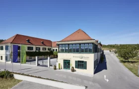 Gebäude der Winzer Krems mit rotem Dach und grünen Fensterläden, umgeben von Weinbergen.