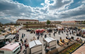 Menschenmenge auf einem Schlossgelände mit Verkaufsständen und Sitzbereichen, umgeben von historischen Gebäuden und blühenden Blumen.