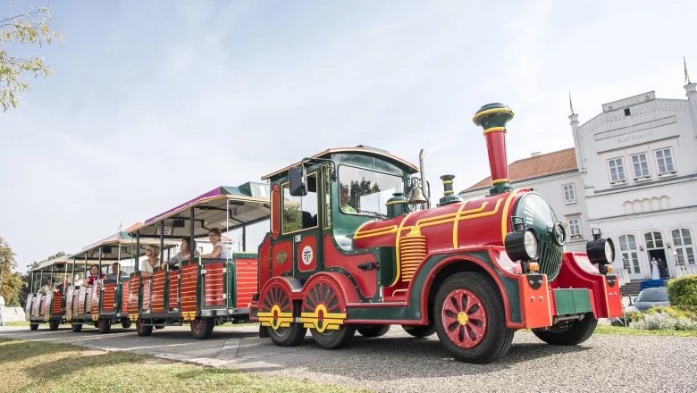 Ein bunter Touristenzug mit mehreren Waggons f&auml;hrt an einem wei&szlig;en Geb&auml;ude vorbei.