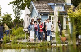 Eine Gruppe von Menschen steht vor einem kleinen blauen Haus mit Terrasse und Sonnenschirmen, umgeben von Pflanzen.