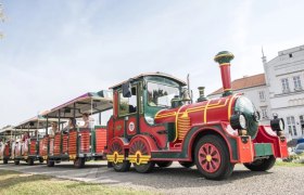Ein bunter Touristenzug mit mehreren Waggons fährt an einem weißen Gebäude vorbei.
